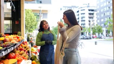 Mutlu genç bayan müşteri olgun kırmızı domatesleri seçiyor ve onları yeniden kullanılabilir bir alışveriş çantasına koyuyor. Bu sırada açık hava pazarında bir manavla iletişim halindeler.