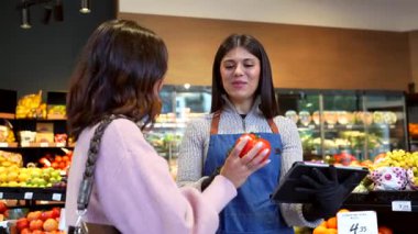 Genç bir kadın müşteriye, modern bir markette ya da yerel markette taze organik domates seçmesinde yardımcı olan bir tabletle arkadaş canlısı bir bayan tezgahtar.