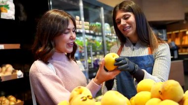 Mavi önlüklü arkadaş canlısı bayan tezgahtar genç bir bayana markette ya da yerel markette olgun, organik sarı meyveleri seçmesinde yardım ediyor.