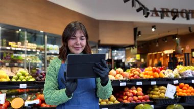 Dost canlısı bir bayan tezgahtar, marketin meyve ve sebze reyonundaki bir müşteriye dijital tablet kullanarak mükemmel hizmet ve modern perakende teknolojisi sunuyor.