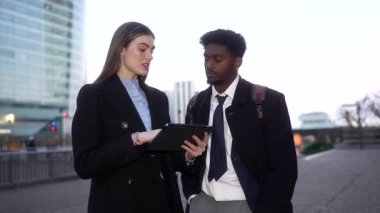 Modern şehir finans bölgesinde gökdelenlerle birlikte yürürken Afrikalı Amerikalı meslektaşına dijital tablette bir proje gösteren profesyonel genç iş kadını.