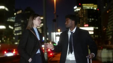 Profesyonel afro-amerikan işadamı ve beyaz iş kadını gece birlikte yürürken, aydınlanmış şehir silüeti ve arka plandaki trafik ile dostça bir sohbete girişiyor.