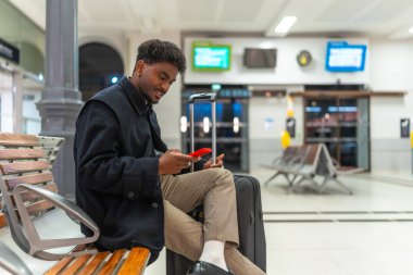 Genç siyah bir adam, tren istasyonundaki ahşap bir bankta rahat bir şekilde oturuyor, kırmızı akıllı telefonunu kullanarak yanında siyah bir bavulla, ulaşımı bekliyor.
