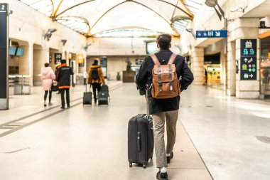 Modern bir tren istasyonunda yürüyen bir iş adamı, bir bavul çekiyor ve sırt çantası takıyor, seyahat, seyahat, iş seyahatleri ve ulaşım kavramlarını vurguluyor.