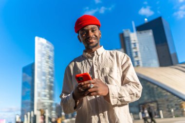 Genç siyahi bir adam kameraya gülümserken açık havada kırmızı bir akıllı telefon kullanıyor. Gündüz boyunca parlak mavi gökyüzünün altında uzun cam binalarla çağdaş bir şehir ortamında duruyor.