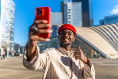 Genç siyahi bir adam kırmızı bir akıllı telefonla selfie çekerken gülümsüyor ve modern mimarinin ve gökdelenlerin önünde mavi gökdelenlerin altında dikilirken baş parmak işareti yapıyor.