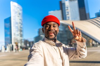Genç siyahi bir adam gülümsüyor ve barış işareti yapıyor. Modern bir şehir şehrinde, parlak mavi gökyüzü altında çağdaş mimarisi olan bir selfie çekerken.