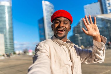 Genç siyahi bir adam selfie çekiyor, geniş bir gülümsemeyle kameraya el sallıyor, açık mavi gökyüzü altında gökdelenleri olan modern bir şehir bölgesinde dostça bir jest yapıyor.