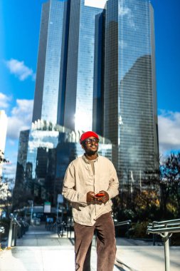 Modern şehir ortamında kendine güvenen genç siyah bir adam şık bir kıyafet ve kırmızı bir bere giyiyor. Gökdelenlerle dolu bir akıllı telefon tutuyor. Parlak mavi gökyüzünü yansıtıyor.