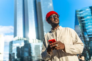 Kırmızı bereli ve gözlüklü genç bir adam, elinde modern gökdelenlerle kırmızı bir akıllı telefon tutarken gülümsüyor ve yukarı bakıyordu.