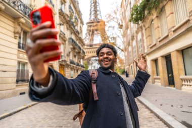 Genç siyahi turist gülümseyip telefonuna selfie kaydediyor, el sallıyor, arka planda ikonik Eyfel kulesi olan bir Paris caddesinde duruyor.