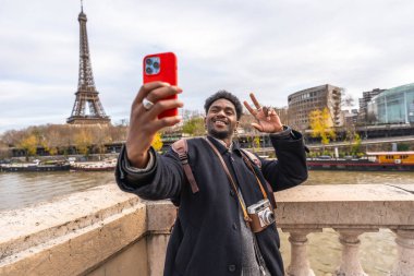 Gülümseyen ve barış işareti yapan genç bir adam akıllı telefonuyla selfie çekerken, arka planda Eyfel kulesi ve Seine nehri ile olan mutlu seyahat deneyimini yakalıyor.