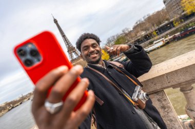 Genç siyahi adam gülümsüyor ve akıllı telefonuyla selfie çekiyor. Paris 'te, arka planda Eyfel kulesi olan şehre tepeden bakan unutulmaz bir seyahat anı yakalıyor.