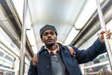 Kışlık montlu ve şapkalı genç bir adam kalabalık bir metroda metro direğini tutuyor. Günlük gidiş gelişler, şehir seyahatleri ve rutin hareketler sırasında dalgın dalgın bakıyor.