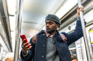 Metrodaki Afrikalı Amerikalı bir adam elinde bir sopayla gezintiye çıkarken akıllı telefonunu karıştırıyor sırt çantası ve odaklanmış ifadesiyle sokak kıyafetleri giyiyor.