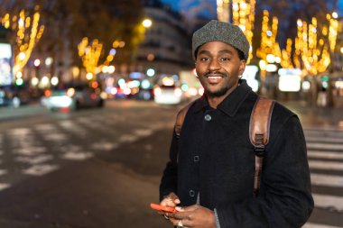 Gece şehir ortamında gülümseyen genç bir adam, elinde bir cep telefonu ve arka planda bulanık araba ışıkları ve dekore edilmiş ağaçlarla bir şehir caddesinde duruyor.