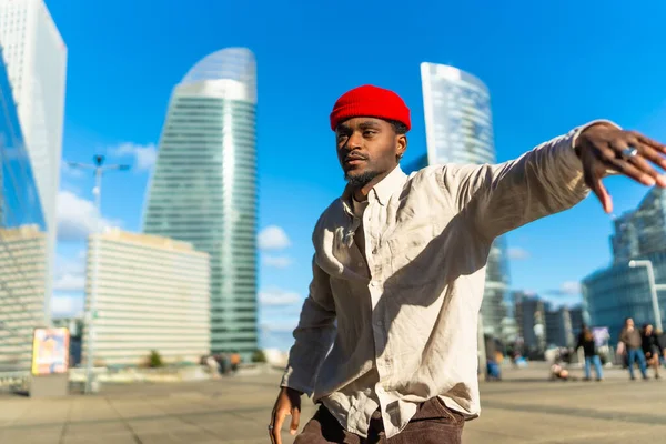 Siyah adam kırmızı bir bere takıyor ve güneşli bir günde arka planda modern cam gökdelenler olan şehir meydanında ritmik bir şekilde hareket ederek neşe ve özgürlüğü ifade ediyor.
