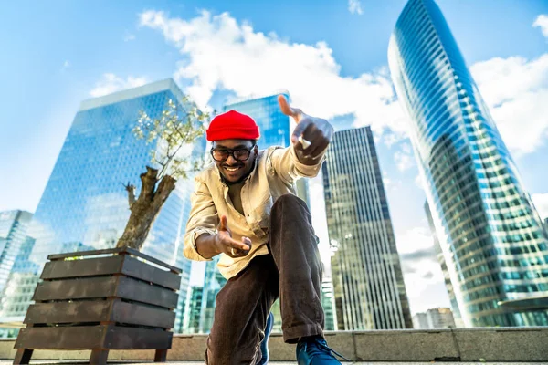 Kırmızı bir bere ve gözlük takan mutlu genç adam kameraya doğru gülümsüyor ve hareket ediyor. Parlak mavi gökdelenlerin altında etkileyici gökdelenleri olan canlı bir şehir ortamında duruyor.