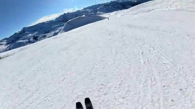 Bir kayakçının geniş, güneşli bir yamaçtan kayak ve direklerle açık mavi bir gökyüzünün altında süzüldüğünü görmek heyecan verici bir bakış açısı.