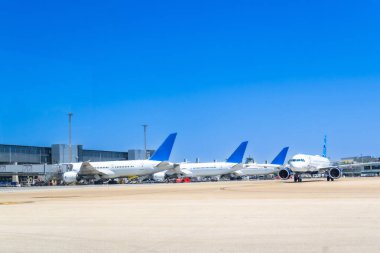 Açık ve güneşli bir günde havaalanı kapılarına park etmiş birkaç beyaz ve mavi uçak bir sonraki kalkış veya gelişlerini bekliyor ve canlı bir havacılık aktivitesi manzarası yaratıyorlar.
