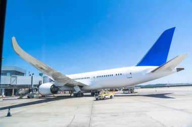 Havaalanı kapısına park etmiş beyaz ve mavi yolcu uçağı, güneşli bir gökyüzü altında kalkışa hazırlanıyor. Açık mavi ufuk, mükemmel bir seyahat gününü aydınlatıyor.