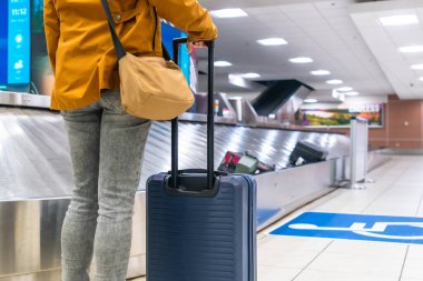 Tourist waiting at the baggage claim area in an airport, holding a suitcase while observing the conveyor belt for arriving luggage after a long journey