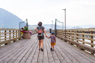 Anne ve oğlu, sakin sular ve somon kolundaki görkemli dağlarla çevrili, doğa ile iç içe geçmiş aile bağlarını kucaklayan, el ele yürüyorlar.