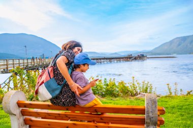 Anne ve oğlu bir bankta oturmuş, somon kolunda Shuswap Gölü ve halka açık rıhtımın manzarasının keyfini çıkarıyorlar. Güneşli bir günde, yemyeşil ve dağlarla çevrili.