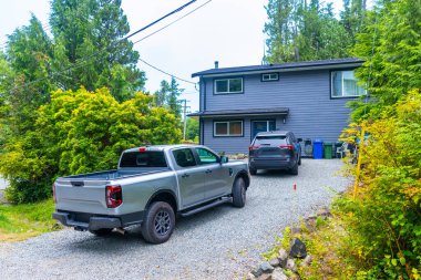 Modern iki katlı gri evde bir kamyonet ve SUV, Tofino 'nun verimli yeşillikleriyle çevrili bir çakıl yola park edilmiş, tipik İngiliz Kolombiya manzarası.