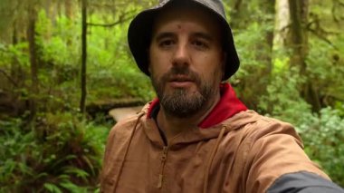 Coşkulu içerik yaratıcısı kova şapka takıyor ve kırmızı kapüşonlu açık kahverengi bir ceket giyiyor. Vancouver Adası 'nın yemyeşil yağmur ormanlarında bir video blogu kaydediyor.