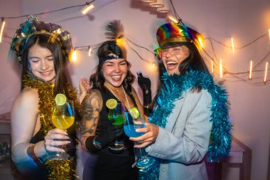 Üç genç bayan arkadaş eğlenceli bir yeni yıl partisinin tadını çıkarıyorlar, renkli kokteyllerle kutluyorlar, kostüm şapkaları giyiyorlar ve birlikte gülerken süslü püslü şeyler giyiyorlar.