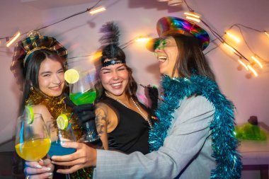 Üç gülümseyen genç arkadaş parti kostümleri ve şenlikli aksesuarlar giyip neşeli içkilerle özel bir olayı kutluyorlar. Neşeli bir gecenin tadını çıkarıyorlar.