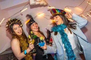 Canlı bir partide, ellerinde renkli kokteyller, şenlikli kostümler ve süslü süslemeler olan üç farklı genç kadın mutlu bir şekilde kutluyor ve dans ediyor.