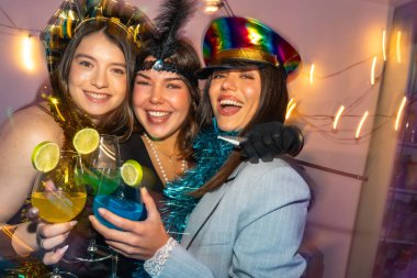 Üç mutlu genç kadın renkli kokteyllerle gülüyor ve kadeh kaldırıyor, kostümler içinde arkadaşlarıyla canlı ve canlı bir atmosferde kutlama yapıyorlar.