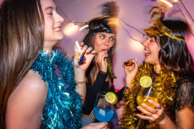Üç mutlu genç kadın canlı bir yeni yıl partisinde ya da kostüm partisinde, şen şakrak aksesuarlar takıyor, parti kornaları çalıyor ve ellerinde parlak renkli içkilerle kokteyller tutuyorlar.