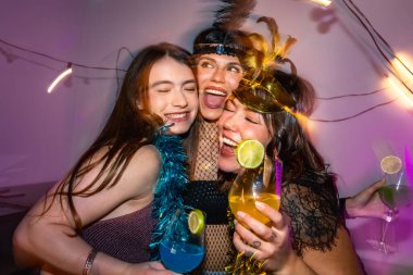 Kokteyller ve kıyafetlerle dolu, renkli parti ışıkları altında arkadaşlık, eğlence ve iletişim saçan, kucaklaşan, gülen ve eğlenen üç mutlu genç kadın.