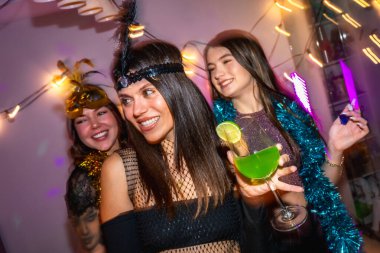 Üç arkadaş gülümsüyor ve evde yeşil bir kokteyl tutuyor ve dekoratif ışıklarla canlı atmosferin tadını çıkarıyor.
