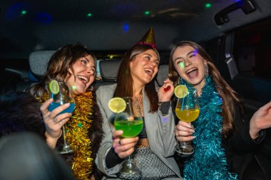 Parti şapkaları ve süslü çelenkler takmış üç arkadaş bir arabanın içinde gülüyor ve renkli kokteyller kızartıyorlar, neşe ve spontane eğlenceyle dolu neşeli bir gecenin tadını çıkarıyorlar.