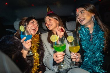 Üç mutlu genç kadın bir arabada seyahat ediyor, birlikte gülüyor ve renkli kokteyller içiyorlar, süslü püslü şeyler giyiyorlar ve bir parti şapkası takıyorlar, neşeli bir gece kutlamasının tadını çıkarıyorlar.