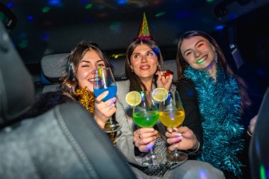 Üç gülümseyen genç kadın neşeli şapkalar ve süsler takıyor, ellerinde renkli kokteyller, disko ışıkları arabanın içini aydınlatırken dışarıda eğleniyorlar.