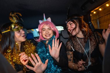 Şenlikli kostümler ve çelenkler giyen üç genç kadın bir gece kutlaması sırasında arabanın arka koltuğunda güvenle seyahat ederken mutlu bir şekilde gülüp dans ediyorlar.