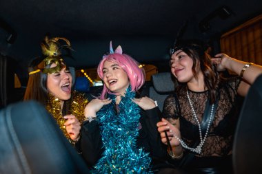 Üç genç kadın gülümsüyor, gülüyor ve eğleniyor. Şenlikli aksesuarlarla kostüm giyip gece bir arabanın arka koltuğunda eğleniyorlar.