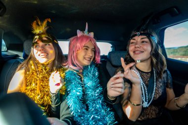 Üç genç kadın bir arabanın arka koltuğunda gülüyor ve kutlama yapıyorlar. Renkli süsler ve şen şakrak aksesuarlar giyiyorlar.