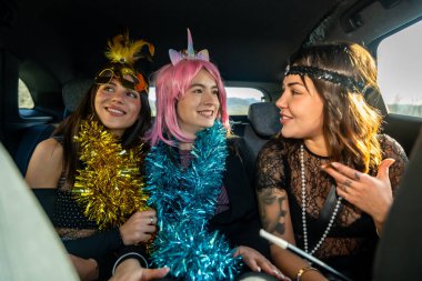 Renkli aksesuarlar ve parti kıyafetleri içinde üç genç kadın bir arabanın arka koltuğunda gülüyor ve konuşuyor, birlikte özel bir olayı kutluyorlar.