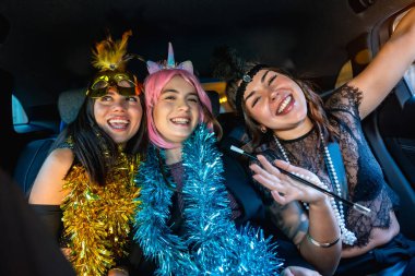 Gülümseyen ve gülen üç genç kadın, çelenk takmış kostümler içinde, arabanın içinde bir partiyi kutluyorlar, gece yolculuk ederken arkadaşlarıyla eğleniyorlar.