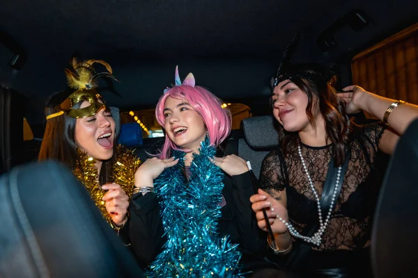 Üç genç kadın gülümsüyor, gülüyor ve eğleniyor. Şenlikli aksesuarlarla kostüm giyip gece bir arabanın arka koltuğunda eğleniyorlar.