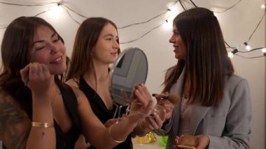 Yılbaşı partisine hazırlanırken evde makyaj yapan üç mutlu genç bayan arkadaş, neşeli ip ışıklarıyla süslenmiş bir odada gülüyor ve sohbet ediyorlar.