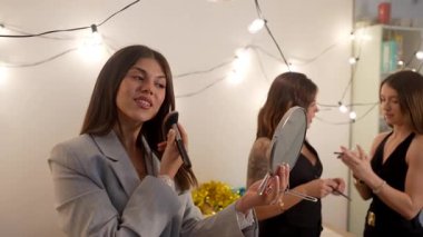 Genç bir kadın makyaj fırçasıyla makyaj tozu sürüyor. Arkadaşları yeni yıl partisi için arka planda hazırlanırken el kamerasıyla aynaya bakıyor.