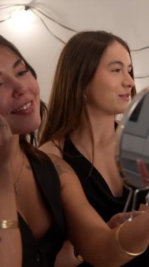 Yeni yıl partisine hazırlanırken birlikte gülümseyen ve eğlenen genç kadınlar, göz farı ve fondöten gibi kozmetik ürünlerini dikkatli bir şekilde kullanırlar.