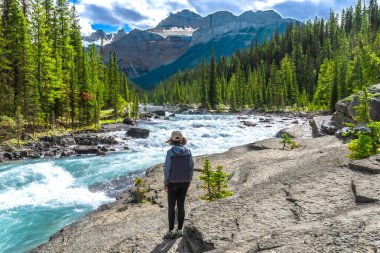 Hataya Nehri 'nin turkuaz sularına hayran olan kadın yürüyüşçü güneşli bir günde Banff Ulusal Parkı' ndaki hataya kanyonu boyunca dağlarla ve kozalaklı ormanlarla çevrili.